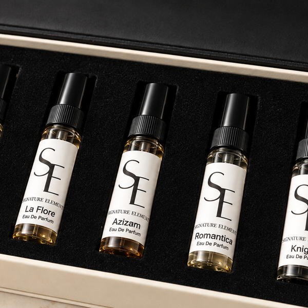 Five small perfume sample vials with black caps and white labels are arranged in a black foam insert inside an open cream-colored gift box. The box lid is open behind the vials, and the set is photographed on a light marble-like surface.