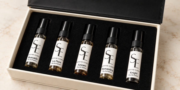 Five small perfume sample vials with black caps and white labels are arranged in a black foam insert inside an open cream-colored gift box. The box lid is open behind the vials, and the set is photographed on a light marble-like surface.